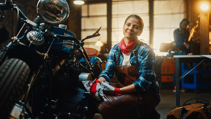 Fototapeta na wymiar Young Beautiful Female Mechanic is Working on a Custom Bobber Motorcycle. Talented Girl Wearing a Checkered Shirt and an Apron. She Smiles at the Camera. Creative Authentic Workshop Garage.