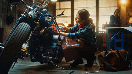 Young Beautiful Female Mechanic is Working on a Custom Bobber Motorcycle. Talented Girl Wearing a Checkered Shirt and an Apron. She Uses a Spanner to Tighten Nut Bolts. Authentic Workshop Garage. © Gorodenkoff
