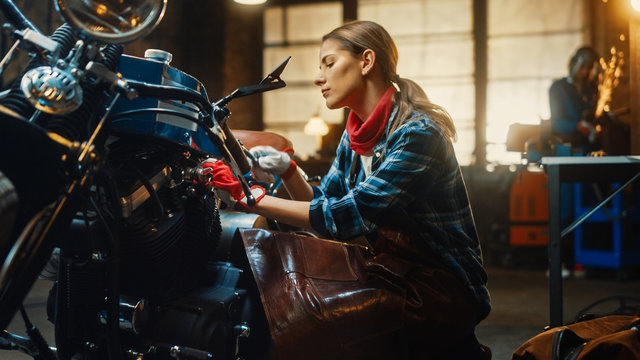 Young Beautiful Female Mechanic is Working on a Custom Bobber Motorcycle. Talented Girl Wearing a Checkered Shirt and an Apron. Creative Authentic Workshop Garage.