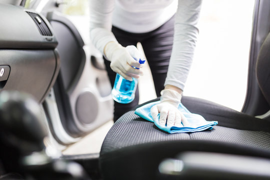Close up of female wearing surgical gloves while cleaning car interior with disinfection liquid sprayer and microfiber cloth.
