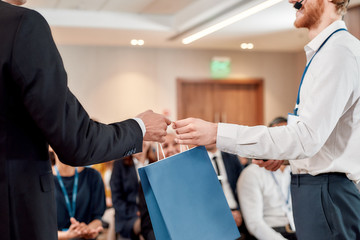 Your reward. Cropped shot of young male speaker receiving gift bag from his male colleague at business meeting, forum