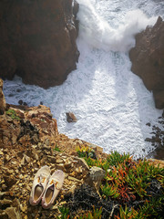 on a sunny day, a pair of old white sneakers stands on a mountain in front of a cliff in the water into the raging ocean. Waves breaking on the rocks. Reminds loneliness.