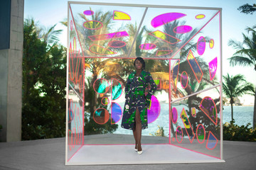 An elegant young black woman with long black hair & beautiful makeup & gorgeous jewelry posing by herself outdoors standing inside a glass box is wearing a colorful coat & hand bag white high heels.