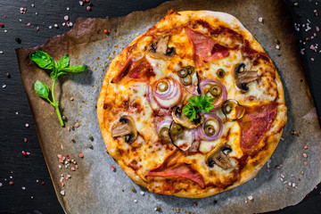 Homemade Italian pizza topped with ham, cheese, onion, mushroom, olive and tomato paste served on a black slate