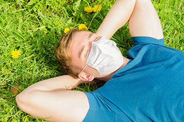 handsome young man in face medical mask  lies on green grass, enjoys resting after self-isolation, quarantine. guy breathes clean air in personal protective equipment for prevention of coronavirus