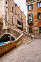 A beautiful stone bridge with a staircase across the Venetian Canal on the streets of Venice, in Italy. The facades of beautiful brick buildings with Venetian-style windows and wooden shutters.