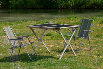 Folding camping chairs and table on the green lawn by the river. Folding garden chairs and table