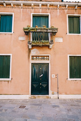 Fototapeta na wymiar Close-ups of building facades in Venice, Italy. Two-storey peach-colored building with green wooden shutters for windows and door. A balcony above the front door with a forged metal grille