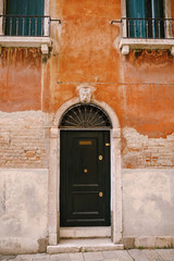 Close-ups of building facades in Venice, Italy. A wooden black door in an arched doorway with a metal grille on top, in the facade of a brick orange building in the Venetian style.