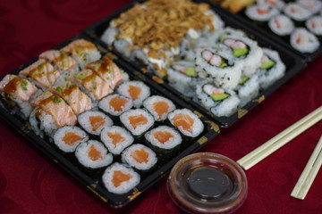 Sushi takeaway box with salmon Hosomaki, Californias, box bowl of soy sauce, hashi chopsticks on red tablecloth