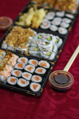 Sushi takeaway box with salmon Hosomaki, Californias, box bowl of soy sauce, hashi chopsticks on red tablecloth, vertical