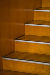 Detail of wooden stairs, a bit worn, with an aluminium leading edge, rising up to the right next to a wooden wall.