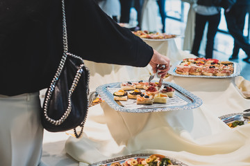 A woman taking a small sandwich from a silver plate on a banquet in a hotel using tongs.