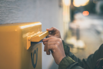 Hands of a white young caucasian woman seen inserting a mail envelope or a letter into a yellow...