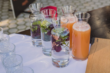 Glass water bottles or jars with water and juice, decorated with flowers on an event or fancy outdoor gala gathering, with fingerfood visible around