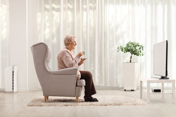 Elderly woman with a cup of coffee seated in an armchair in front of tv