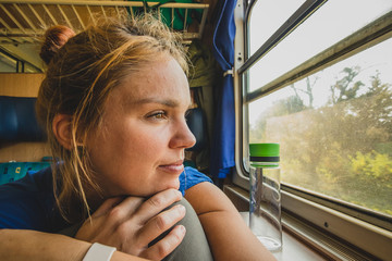 Caucasian woman using looking through a window of an older train with vintage seats and wooden walls. Woman commuting with an older retro train. Sun flare visible