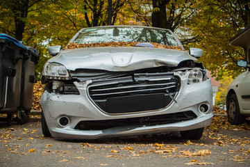 Frontal part of a small city vehicle smashed. Demolished and crashed front of a silver car, parked on a public parking space between the trees and trash cans.