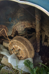 Heavily rusted drum brakes or exterior of brake drum with coil spring and rear axle on an abandoned vintage vehicle. A wheel is missing on an old blue car.