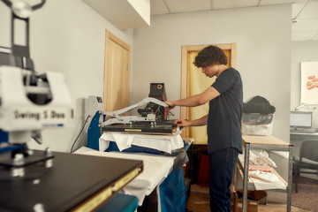 Crafting process. Young cheerful male worker using heat press transfer machine for printing t-shirt...