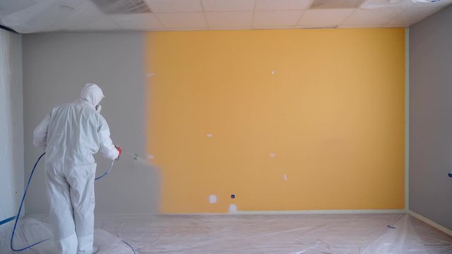 Worker wearing white workwear painting the orange wall in white color by airless spray gun. Airless Spray Painting. Repair at home or office.