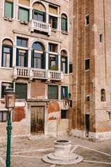 Close-ups of building facades in Venice, Italy. An old street well in the square in front of a brick house. There are many Venetian-style windows on facade of building. Vintage street lamp is green.