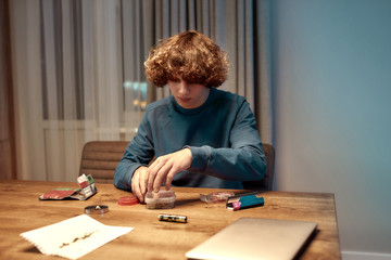 Time to prepare. Young man or teenager making joint, cannabis cigarette sitting at home in the kitchen. Marijuana tools on the table. Cannabis and weed legalization concept