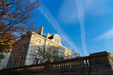 A white mansion pictured in front of the blue sky with two condensation trails, with a tree with orange leaves next to it