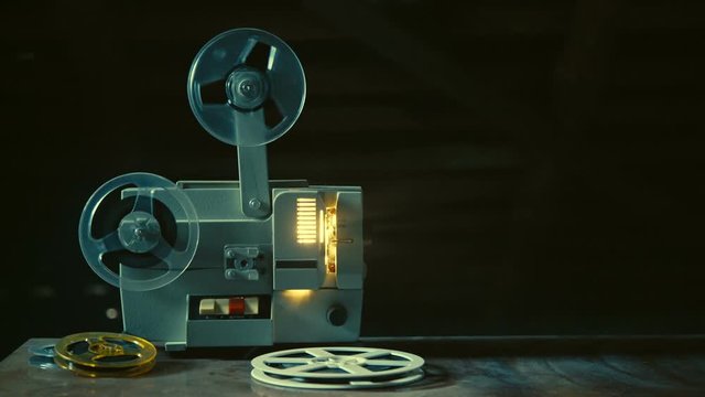 Vintage movie projector is standing on table, switched on, film reels are rotating, lamp is burning inside, several film reels are lying besides, creating magic atmosphere of old cinema, Slow motion.