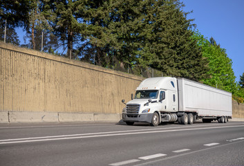 Big rig bonnet long haul semi truck transporting goods in dry van semi trailer running on the wide highway with concrete wall on the side