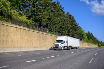 Big rig white semi truck with dry van semi trailer running on the wide multiline highway road with concrete wall and trees on the side