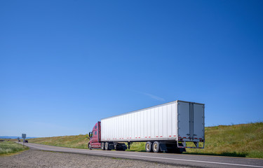 Obraz premium Dark red big rig semi truck transporting commercial cargo in dry van semi trailer driving on the narrow road with hill on the side