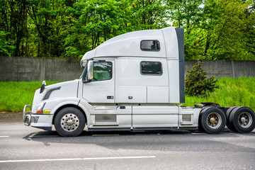 Powerful big rig semi truck tractor running on the road with fence and trees on the side to warehouse for pick up the loaded semi trailer for the next route