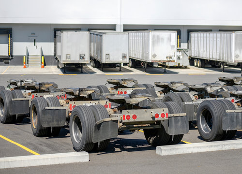 Row of trolleys with axles and fifth wheel tow hitch for semi trucks trains standing on the warehouse parking lot with semi trailers standing in dock gates loading cargo