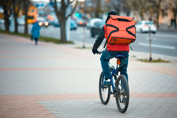 Courier on bicycle deliver an order from the fast food restaurant. Delivery boy on bicycle delivering food, takeaway. Man delivering food with red backpack, riding bicycle trough city street