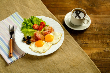 On a wooden table on a linen napkin, a plate with fried eggs with bacon and a cup of coffee.