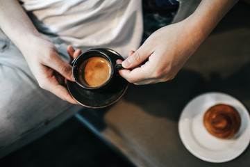 Man in white t-shirt hand holding a hot espresso in black ceramic cup and fresh homemade French croissant on wooden table. Breakfast with coffee and bread.Baked goods in traditional bakery.