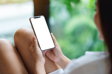 Mockup image of a woman sitting and holding mobile phone with blank white desktop screen , blurred green nature background