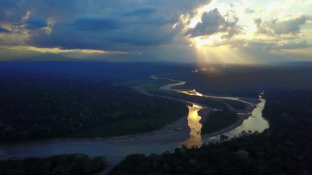 Drone video of Amazon rainforest in Bolivia during sunset. A flowing river and thick jungle are visible with a colorful sunset and mountains in the distance with sun filtering through thick clouds.