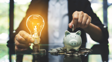 A businesswoman putting coin into piggy bank and a light bulb over coins stack on the table for saving money and financial concept - obrazy, fototapety, plakaty