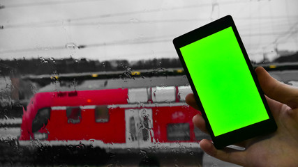 Green screen. A man holds a phone in his hand with a green screen against the background of a window wet from the rain and a commuter train with passengers. Concept of trips and relocations. Template.