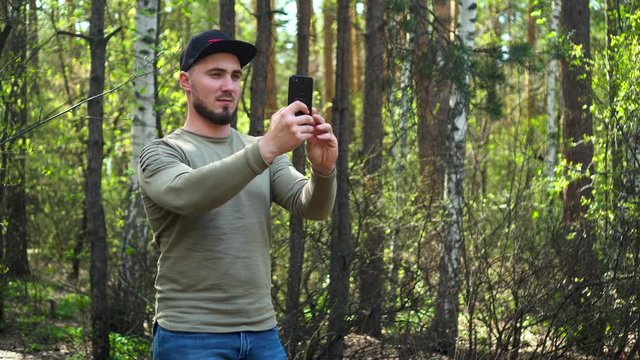 Attractive man in a baseball cap with a beard in the middle of a beautiful forest takes pictures and shoots on a smartphone. Travels through the woods.