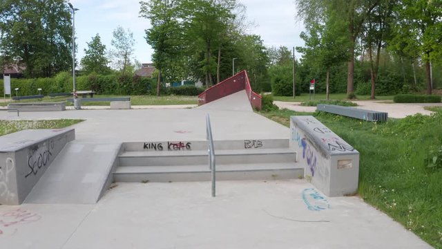 Parallax shot of a small stair set with a small rail and ledge in a concrete skatepark. Green trees in the background. Nobody in the park due to corona crisis.