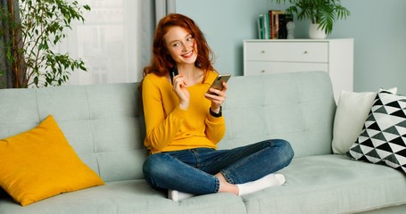 Young beautiful red-haired woman sitting on couch in living room and shopping online on mobile phone.