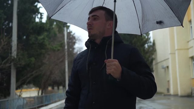 A serious young attractive man stands on a city street with an umbrella in his hands. Rainy weather.