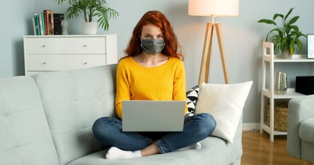 Cheerful young redhead girl in mask sitting on sofa in living room at home and using silver laptop.