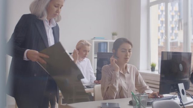 Pan shot of diverse female hotline receptionists sitting at computers and consulting customers. Asian aged woman wearing pantsuit giving chart to mixed-race female operator