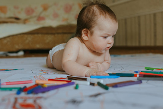 Cute multiracial small girl lying on the floor Pencils crayons and markers are scattered next to the child.
