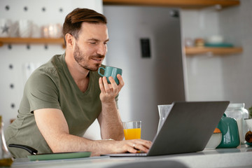 Handsome man drinking coffee in kitchen. Young man reading the news online.