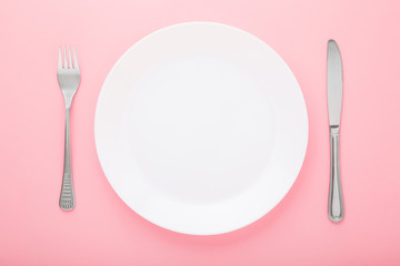 White plate with fork and knife on light pink table background. Pastel color. Closeup. Meal waiting concept. Top view.